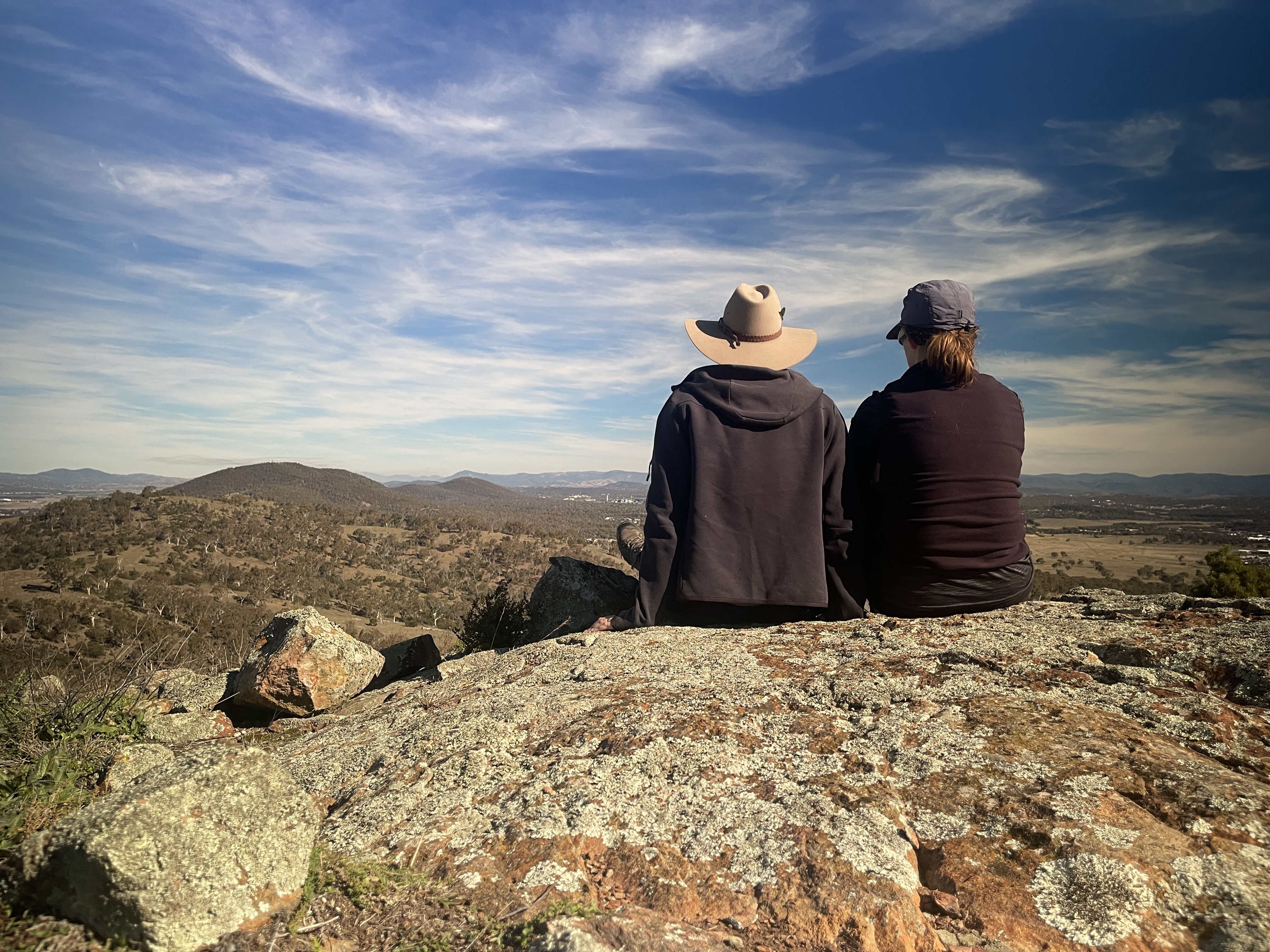 A couple sites on a rock overlooking the Canberra landscape. The man is wearing an Akubra wide brimmed hat and black hoodie and the women is wearing a cap and a black longsleeve top.
